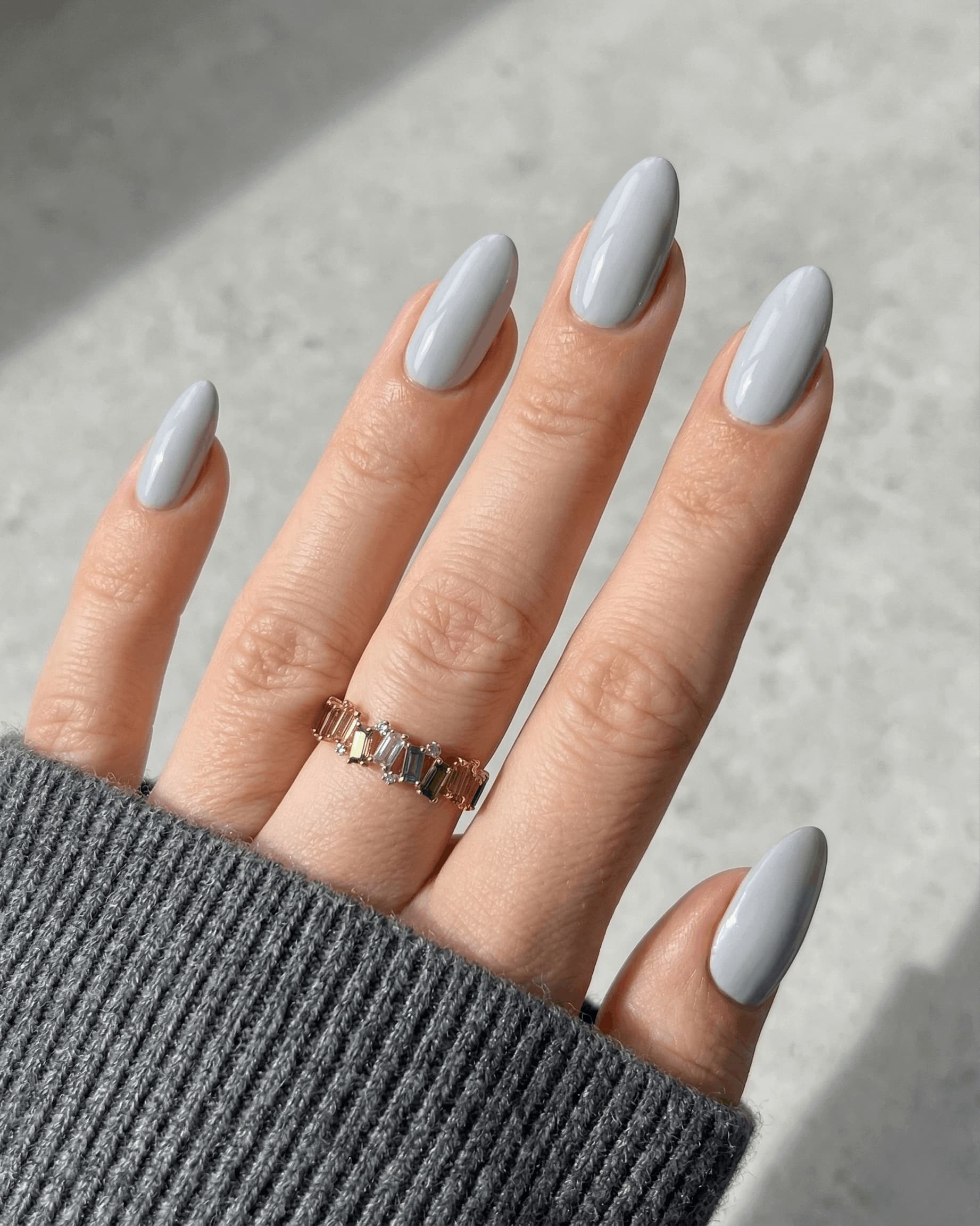 Hand with light gray nail polish wearing a gray sleeve against a neutral background