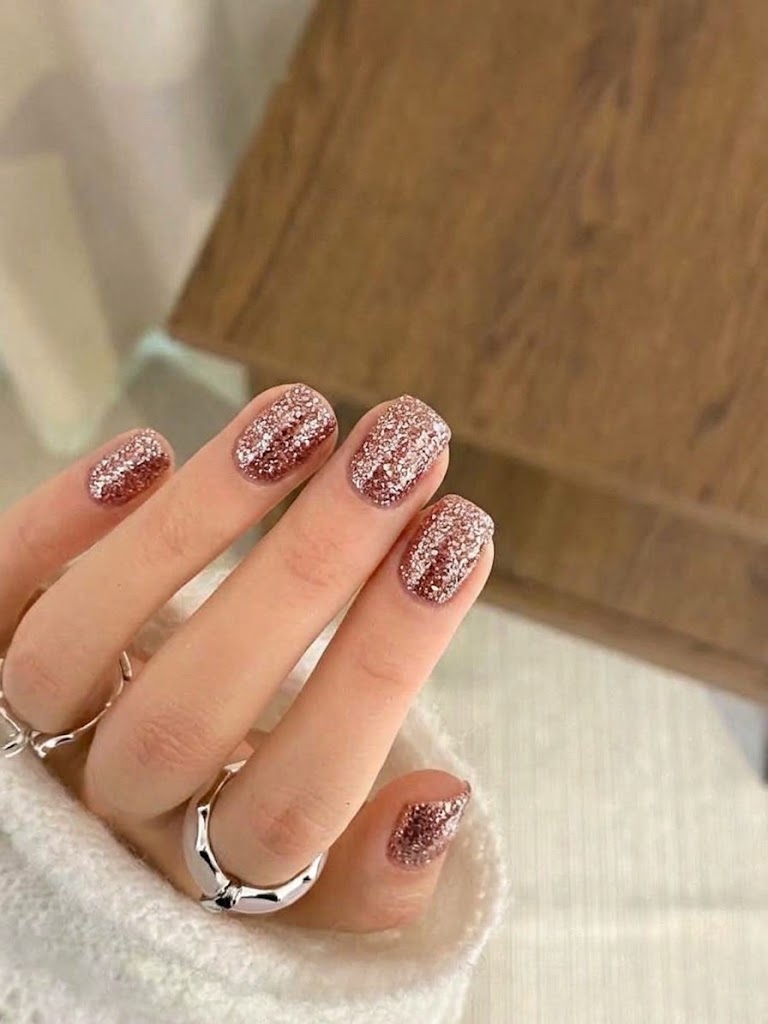 A close-up of a hand with glittery rose gold manicured nails, wearing silver rings and a white knit sleeve, set against a blurred background of a wooden surface and light fabric.