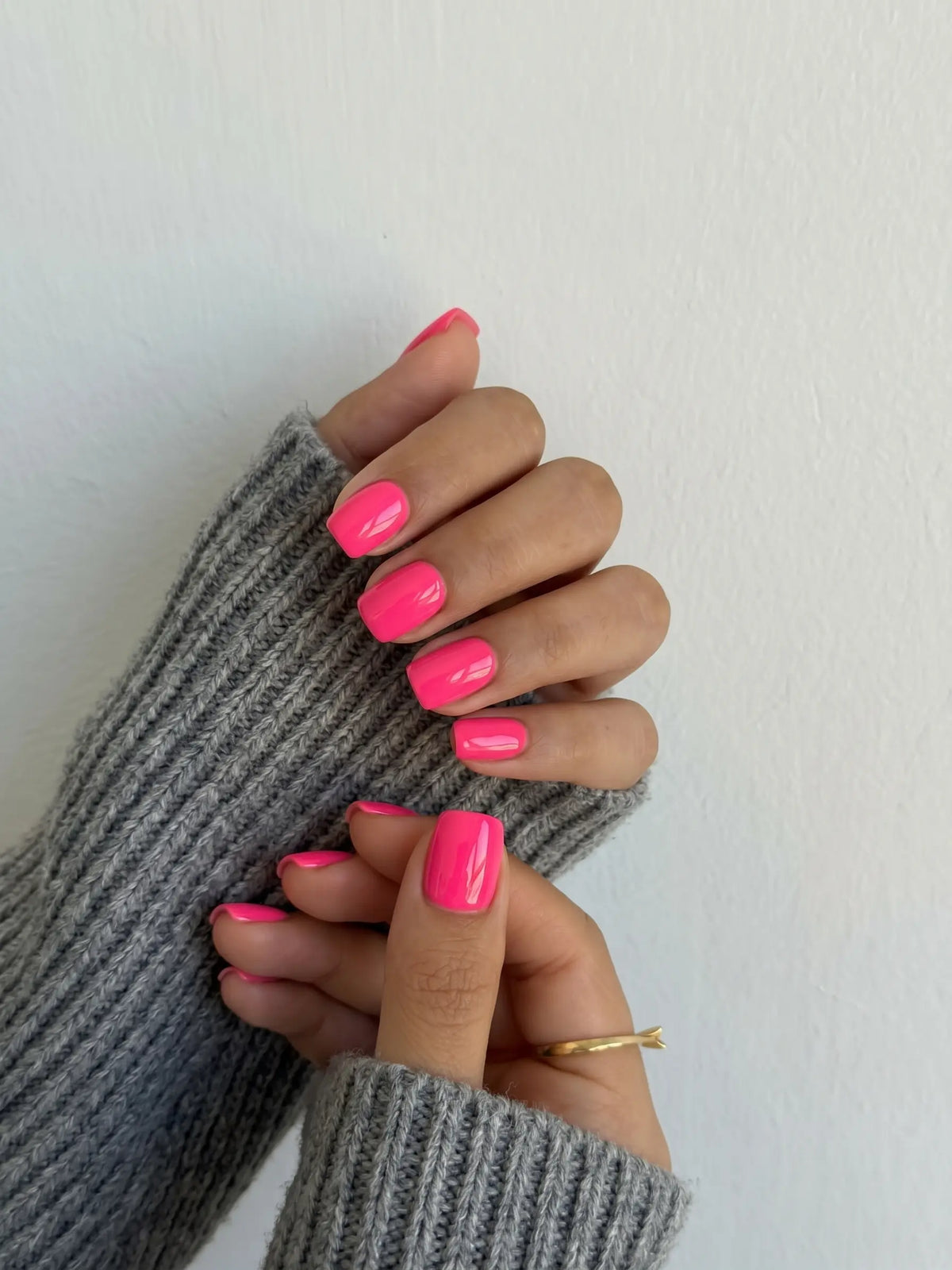 A close-up of hands with bright hot pink manicured nails, wearing a gray ribbed knit sweater against a plain off-white wall.