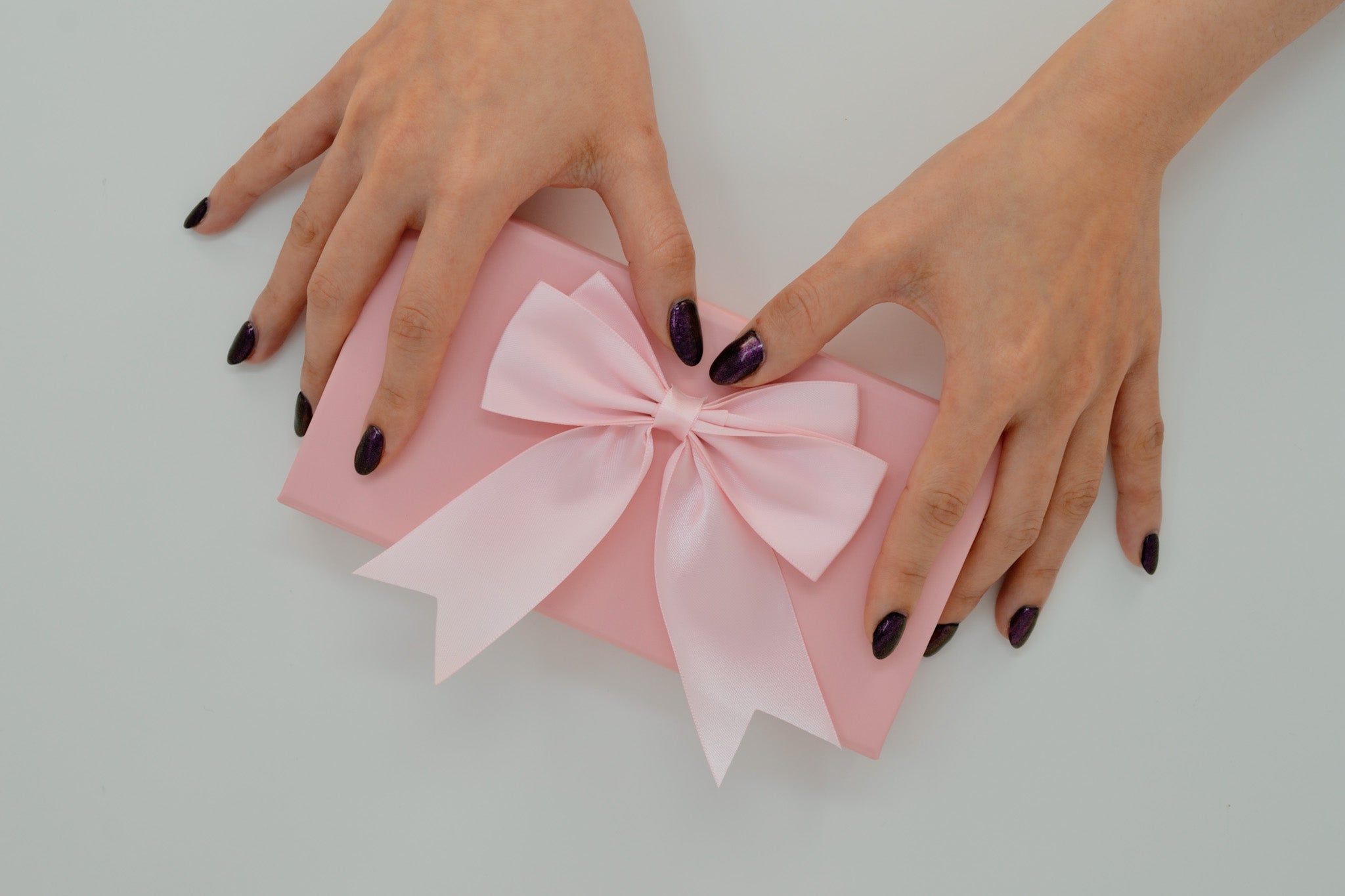 Hands with dark gel nails holding a pink gift box with a satin pink bow